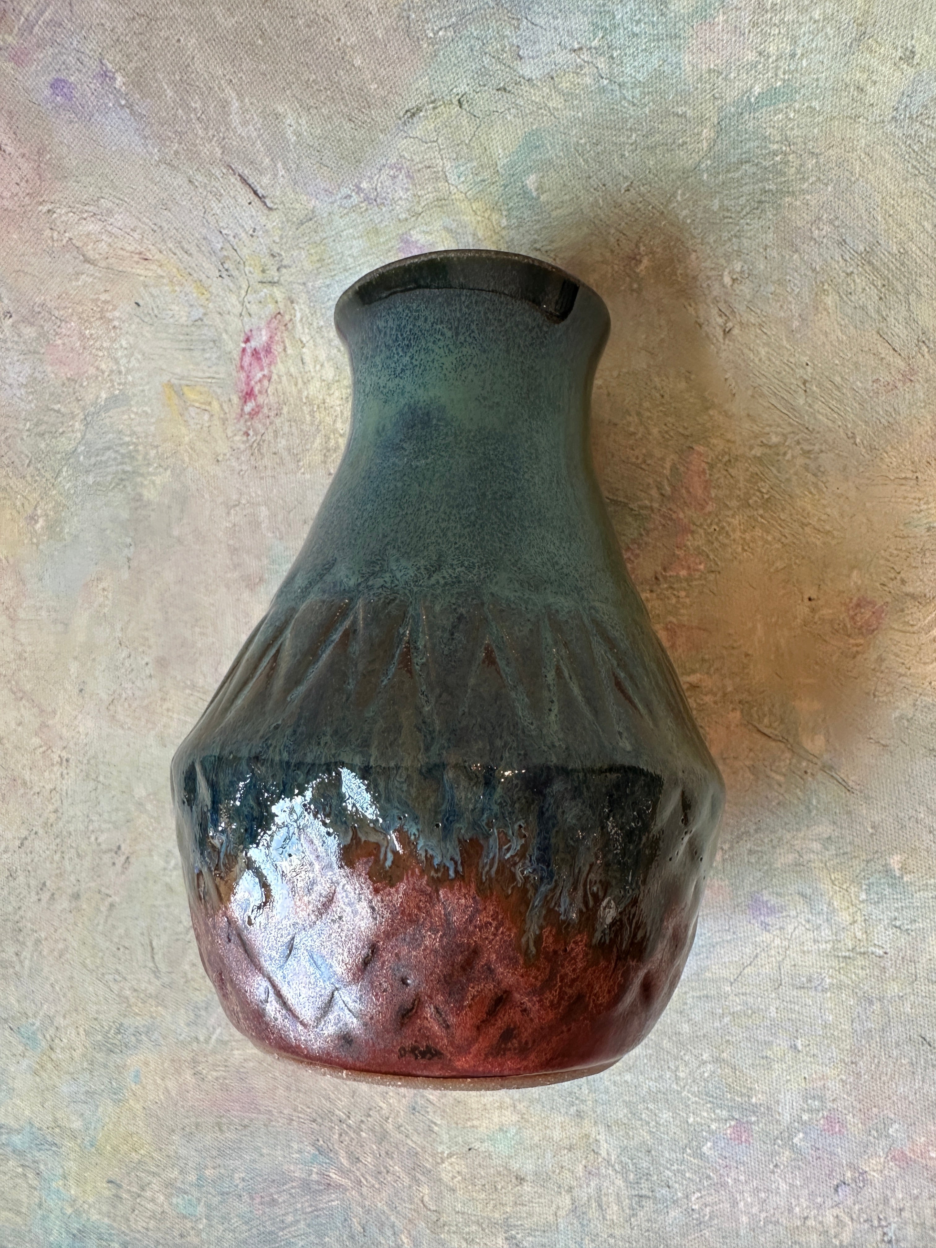 A handmade ceramic vase with a textured, ribbed body and a glossy finish, featuring a dark gray-green glaze on the upper half and a copper glaze on the lower half, resting on a carved textured surface.