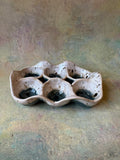 A handmade ceramic six-compartment egg tray with a wavy, organic shape, featuring a speckled beige and white glaze with dark brown/black accents inside the compartments, set against a Blue background.