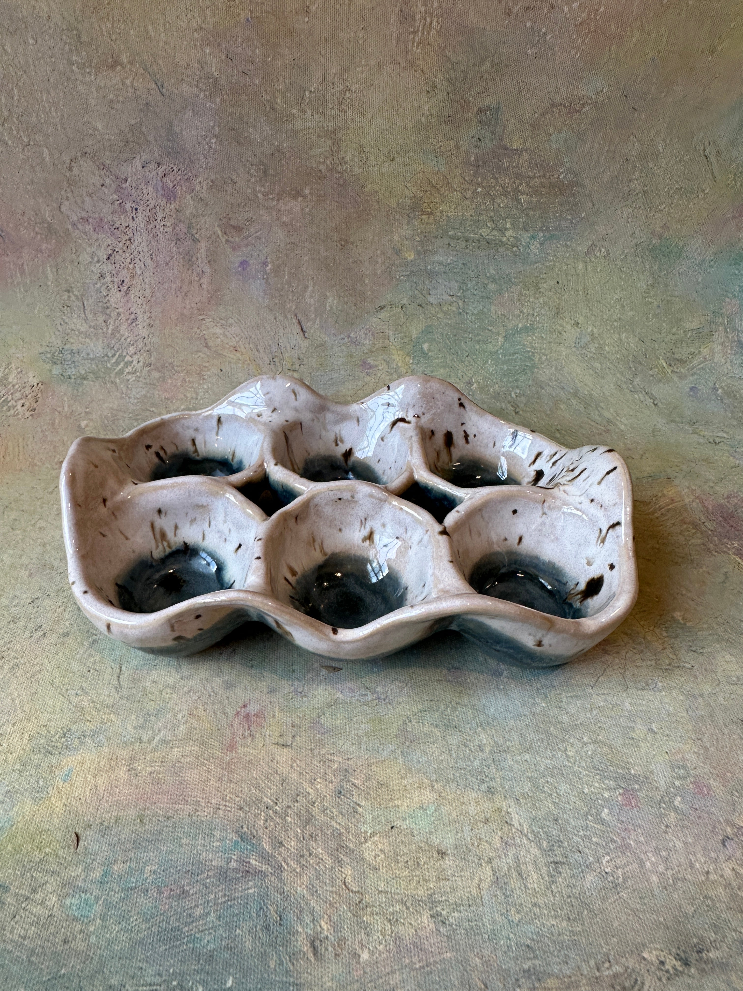 A handmade ceramic six-compartment egg tray with a wavy, organic shape, featuring a speckled beige and white glaze with dark brown/black accents inside the compartments, set against a Blue background.