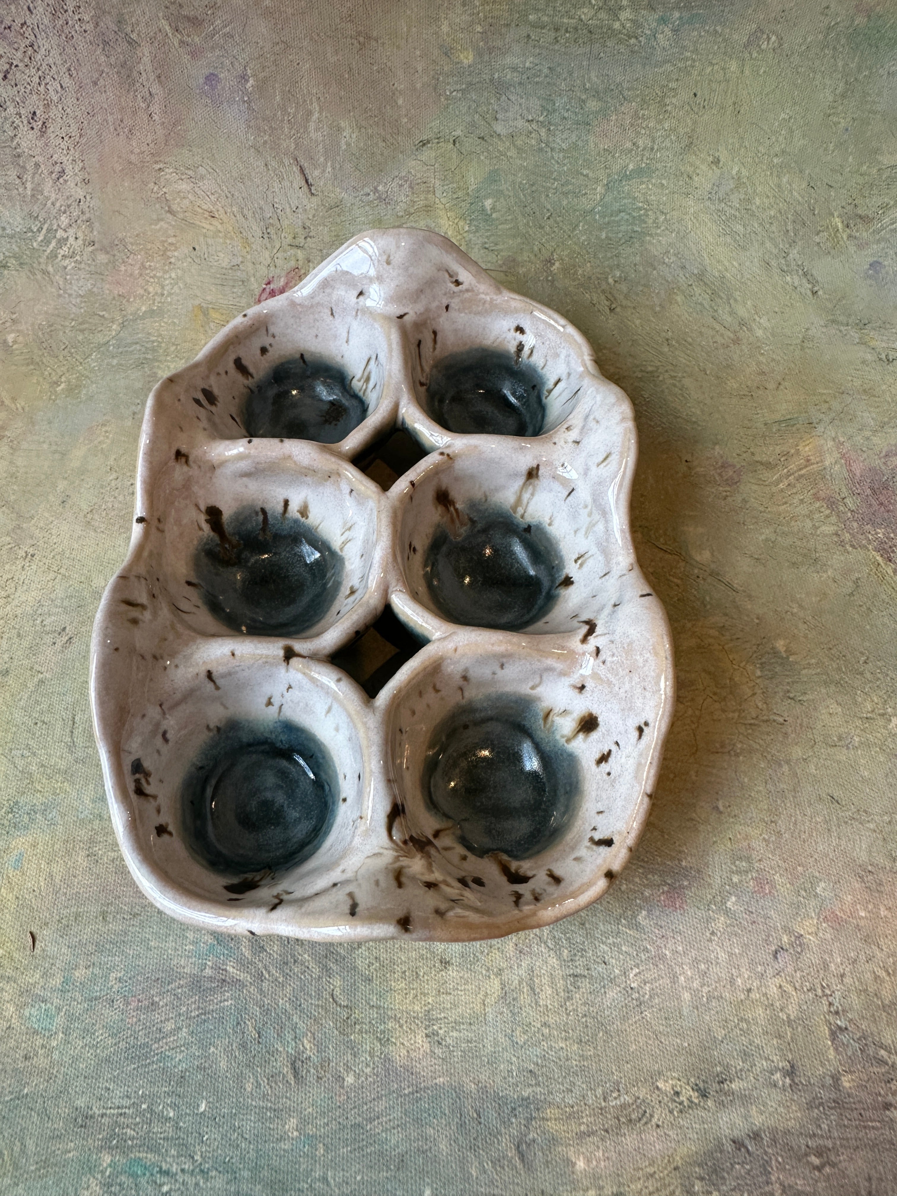 A handmade ceramic egg carton dish with six compartments, featuring a speckled off-white glaze and dark blue centers in each cup, set against a speckled creamy white  background.