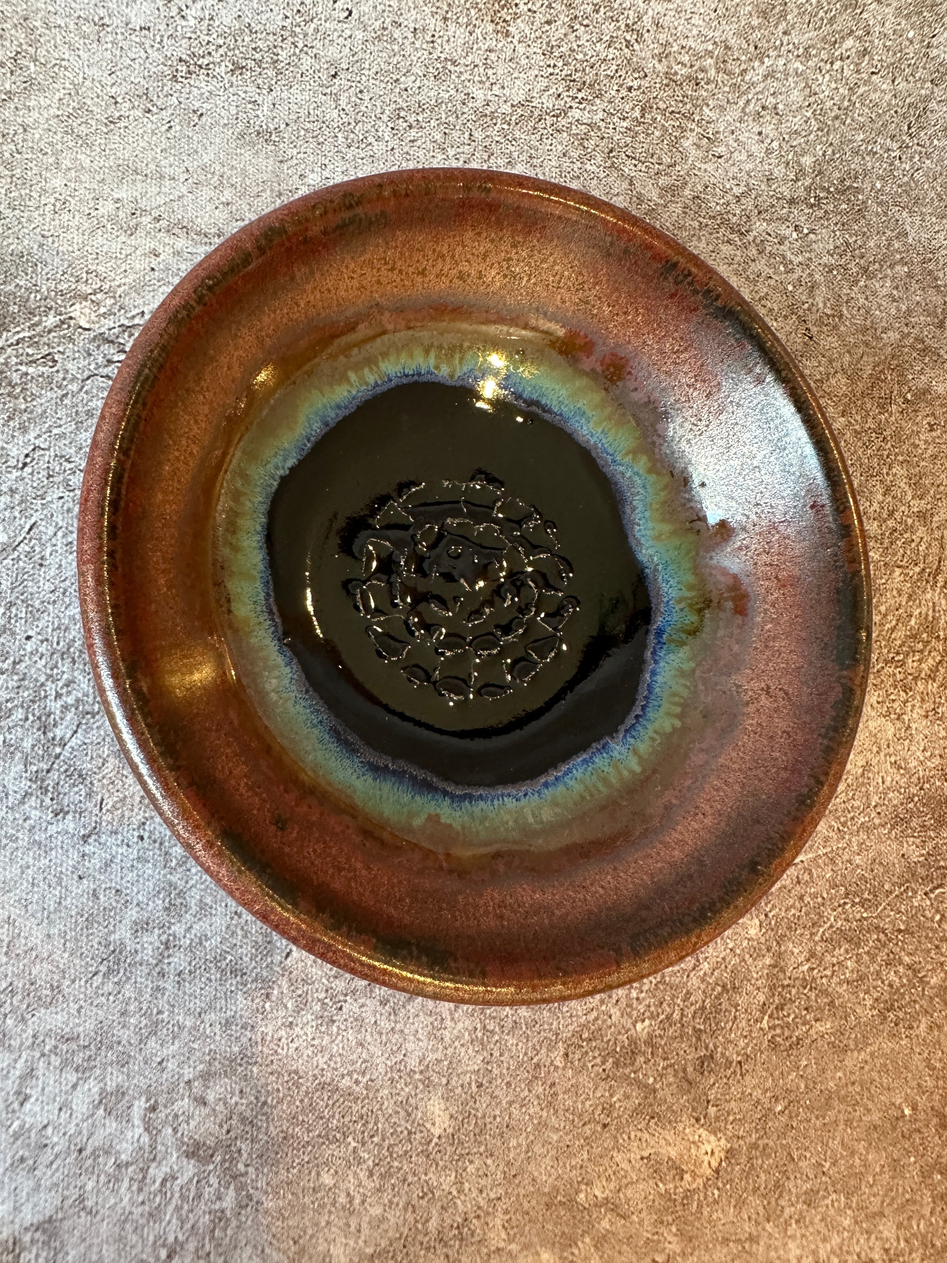A handmade ceramic garlic grater bread dipping bowl with a glossy copper and dark blue black glaze, featuring a blue-green iridescent ring around the center textured surface. Shown from a top-down view.